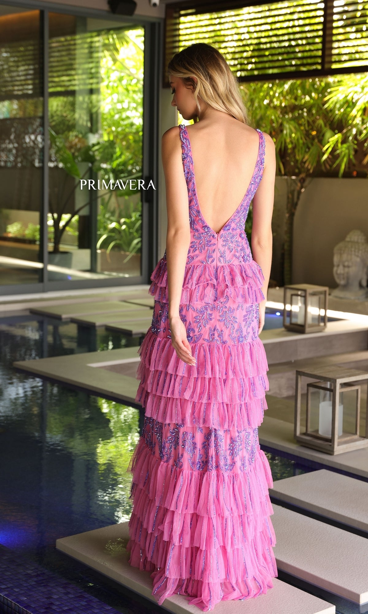 A woman in the Primavera Beaded Long Ruffled Prom Dress 4106 stands near an indoor pool surrounded by lush greenery and lanterns.